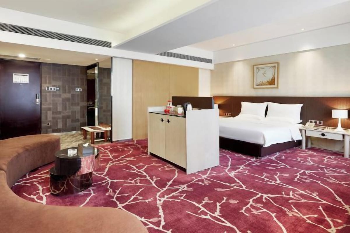 A hotel room with a bed, a desk, a chair, a minibar, and a patterned carpet.