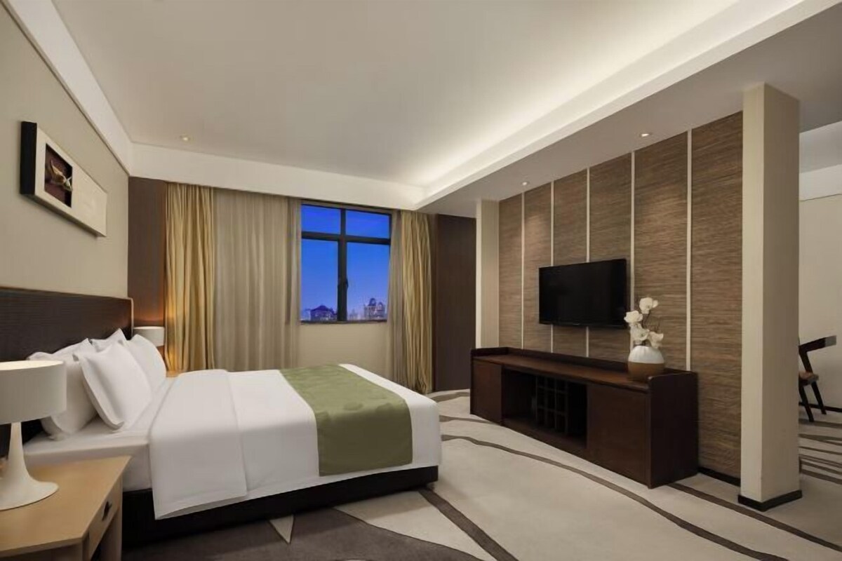 A modern hotel room with a bed, a TV, a desk, and a window with curtains.