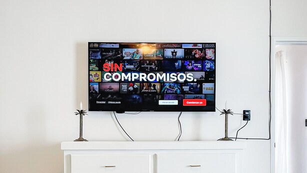 Quarto de hotel com televisão de tela plana na parede, exibindo interface de serviço de streaming, cômoda branca com gavetas e duas camas com colchas verdes.