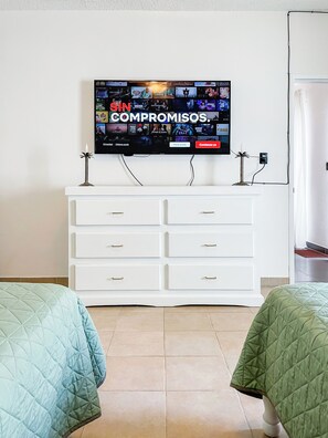 A hotel room with a flat-screen TV mounted on the wall displaying a streaming service interface, a white dresser with drawers, and two beds with green quilts.