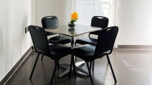 Uma pequena área de refeições com uma mesa de vidro e quatro cadeiras pretas, um vaso com flores amarelas e uma cortina branca.