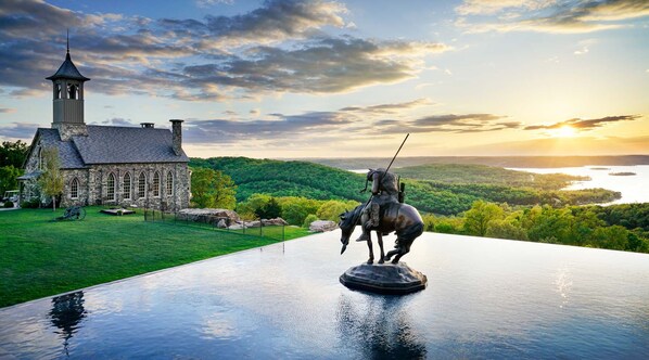 A stone church with a bell tower, a statue of a mounted figure, and a scenic view of a lake and forest.