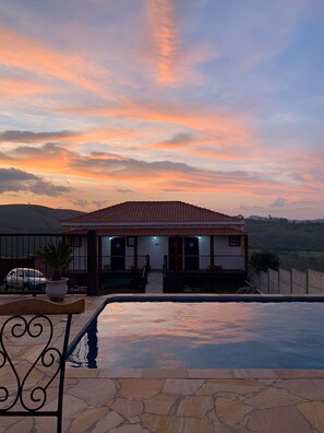 Une maison avec une piscine, entourée d’une clôture, et un coucher de soleil en arrière-plan.