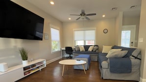A living room with a flat-screen TV, a ceiling fan, a sofa, a coffee table, and a bookshelf. - Waterfront Escape! NYC Ferry, Beach, Modern New Construction 3 bedroom 3 bath (Highlands)