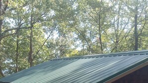 A small, single-story building with a green metal roof and brick walls, surrounded by gravel and trees. - HICKORY HIDEOUT
The cabin is surrounded by a large area of trees for privacy (Mena)