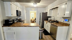 A compact kitchen with white cabinetry, a central island, stainless steel appliances, and a small trash can. - Memphis Retreat w/ Sunroom, Pool Table, Retro Gaming, Gym & Relaxing Vibe (Memphis)