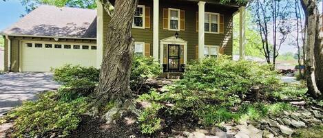 A two-story house with a garage, surrounded by a stone pathway and dense shrubbery.