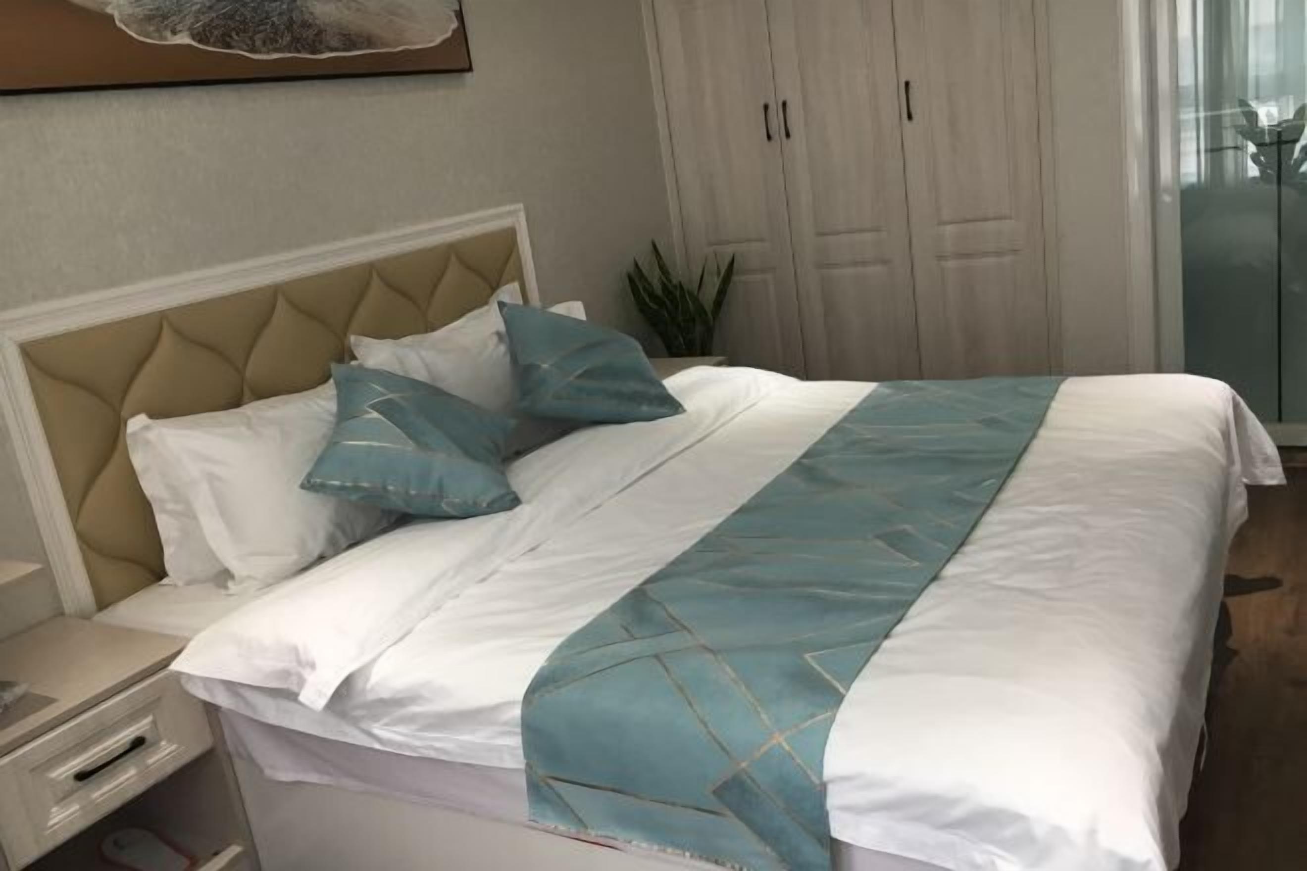 Ein ordentlich bezogenes Bett mit weiß-blauer Bettdecke, einem beige gefärbten Kopfteil und einem Nachttisch mit Lampe.