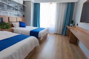 Una habitación de hotel con dos camas, un escritorio, una planta y un amplio ventanal con cortinas.