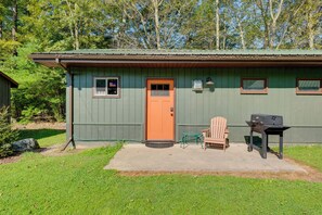 Een kleine, gelijkvloerse hut met een groene buitenkant en een oranje deur. Er staat een houten stoel en een barbecue buiten.