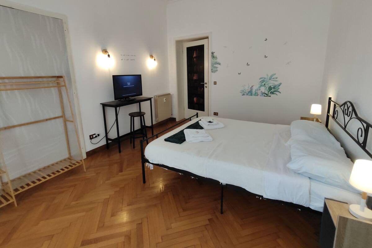 A hotel room with a bed, a desk with a computer, a chair, a radiator, and a door.
