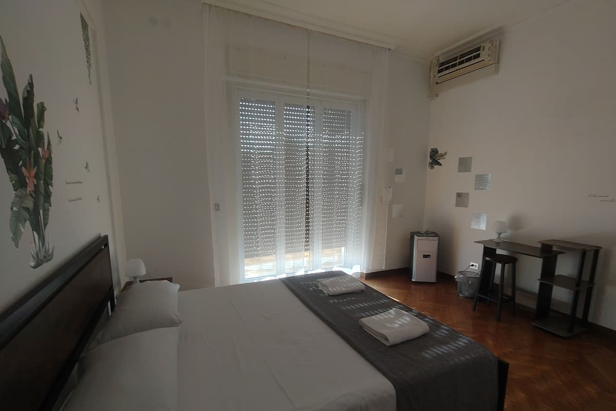 A hotel room with a bed, a desk, a chair, a trash bin, and a window with blinds.