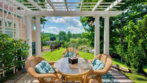 A covered outdoor seating area with wicker furniture and a glass table, overlooking a lush garden and a clear sky. - Mountain Views: Beautiful Nestlenook Farm Home! (Jackson)