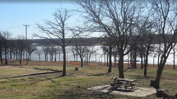 A park with a picnic table and benches, surrounded by trees and a body of water. - Cozy 2-bedroom cabin in front of the lake!!! (Mannford)