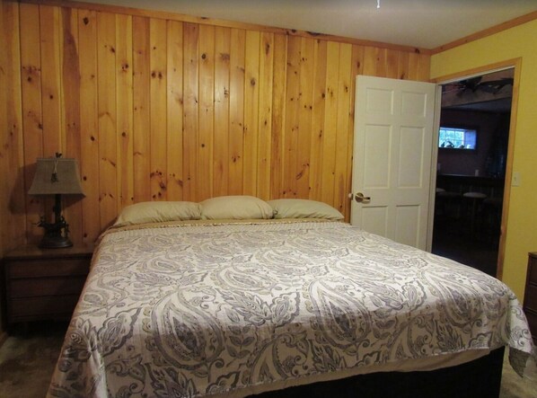 Un dormitorio con pared revestida de madera, una cama con ropa de cama estampada, una mesita de noche con lámpara y una puerta que da a una zona de bar.