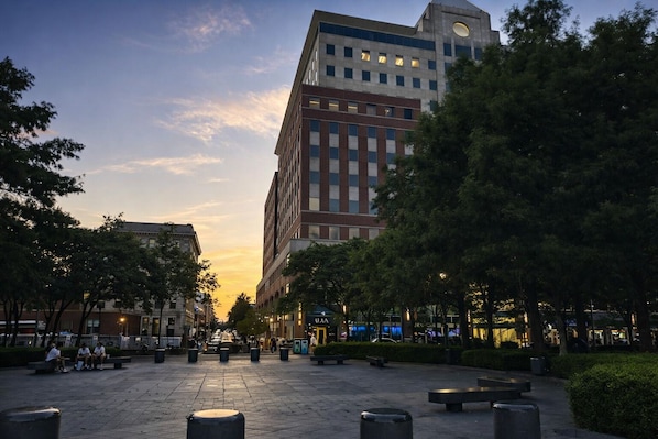 A public square with benches, trees, and a tall building with a circular feature on top.