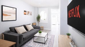 A modern living room with a grey sofa, a coffee table, and a large wall-mounted TV displaying the Netflix logo. - 𖡡 First Street Flat - 1BR Apt just 15 Min to NYC (Hoboken)