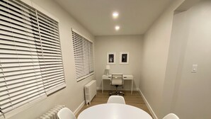 Una habitació amb una taula rodona blanca envoltada de cadires blanques, un radiador i una finestra amb persianes.