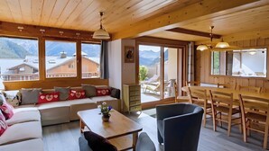 Interior de madera con sofá, mesa de comedor, sillas y ventanas amplias que ofrecen vistas a la montaña.