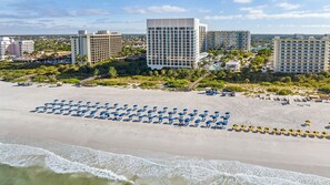 Ein Strand mit Reihen blauer und gelber Sonnenschirme, ein großes Gebäude mit rasterförmiger Fassade und weitere Wohnhäuser im Hintergrund.