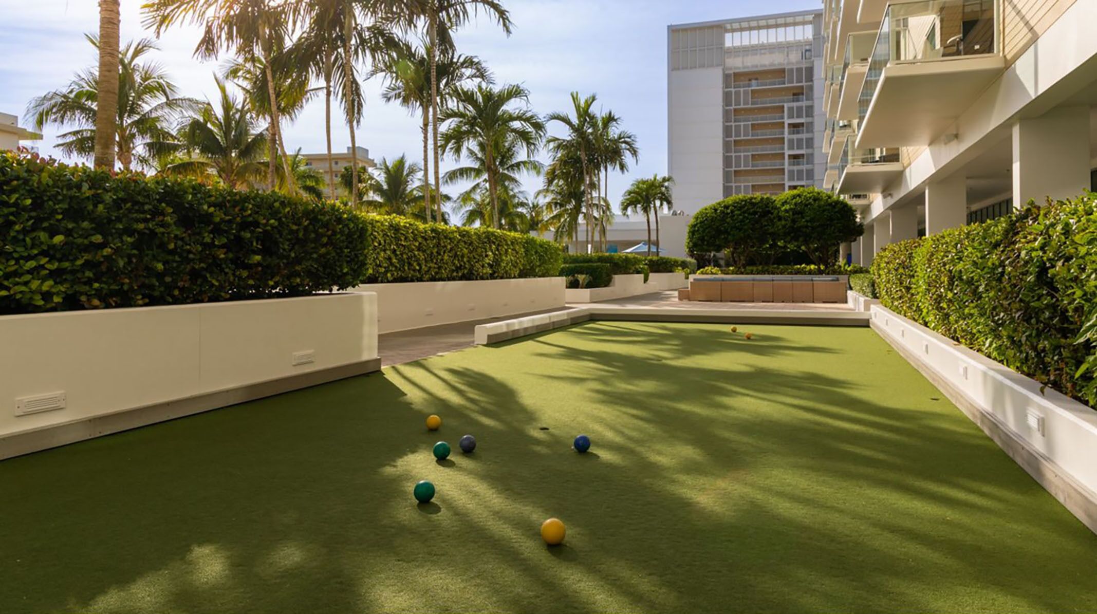 A courtyard with a croquet field, palm trees, and modern buildings in the background.