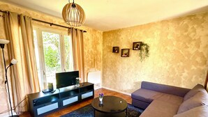 A living room with a sofa, a coffee table, a TV on a stand, a lamp, and a window with curtains. - Vacation House in Quiet Village, Welcome to our 3 bedroom countryside retreet (MONNERVILLE)