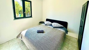 A bedroom with a bed, pillows, a bedside table, a hanging light fixture, and a window with a view of greenery. - Vacation House in Quiet Village, Welcome to our 3 bedroom countryside retreet (MONNERVILLE)