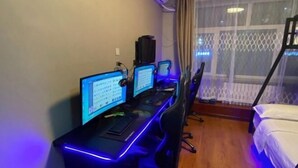 Habitación de hotel con escritorio que incluye tres monitores, un teclado y un ratón. Posee una cama con ropa blanca y una ventana con vista a un edificio.