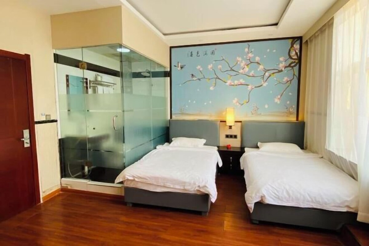 A hotel room with two beds, a glass shower enclosure, a wall-mounted artwork, and a bedside table with a lamp.