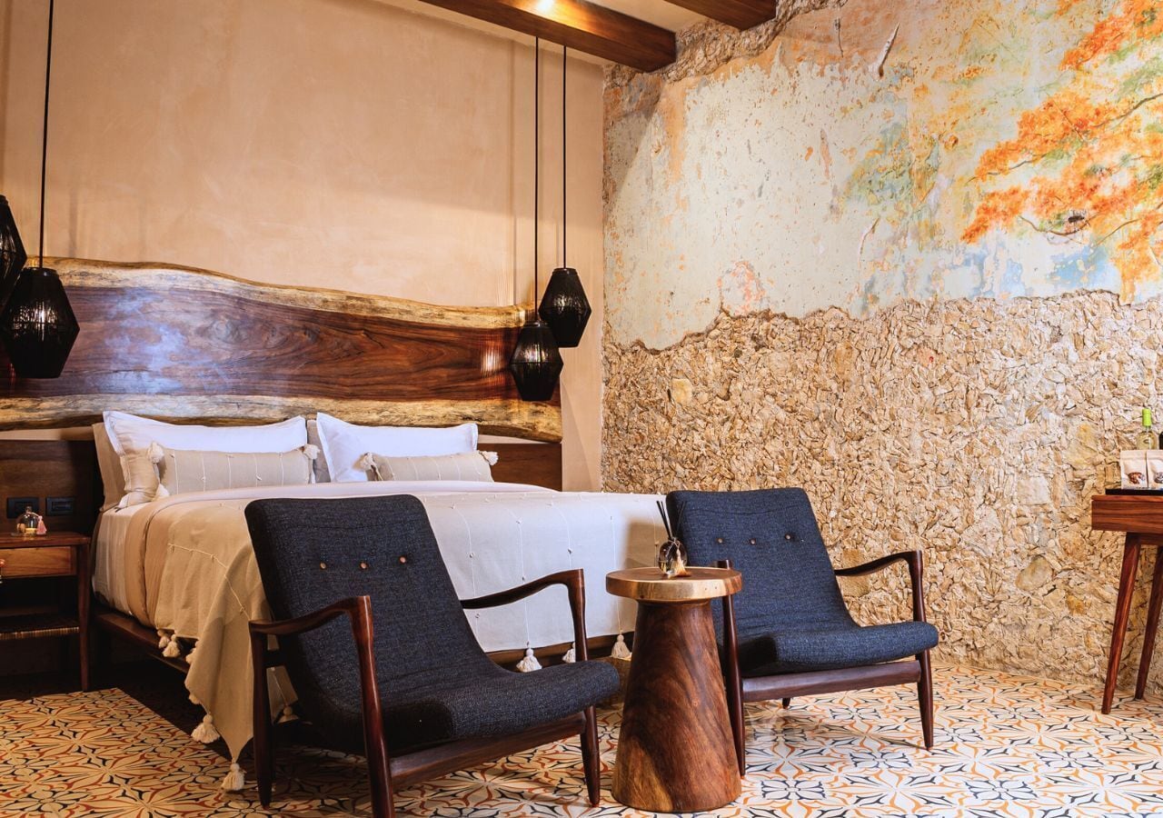 A hotel room with a large bed, two armchairs, a wooden side table, and a stone wall with a textured mural.