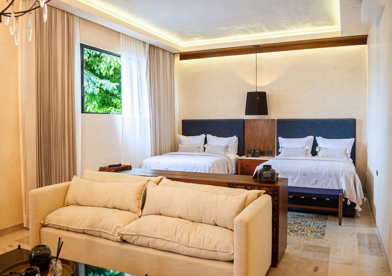 A hotel room with a sofa, two beds, a nightstand, a lamp, and a window with curtains.