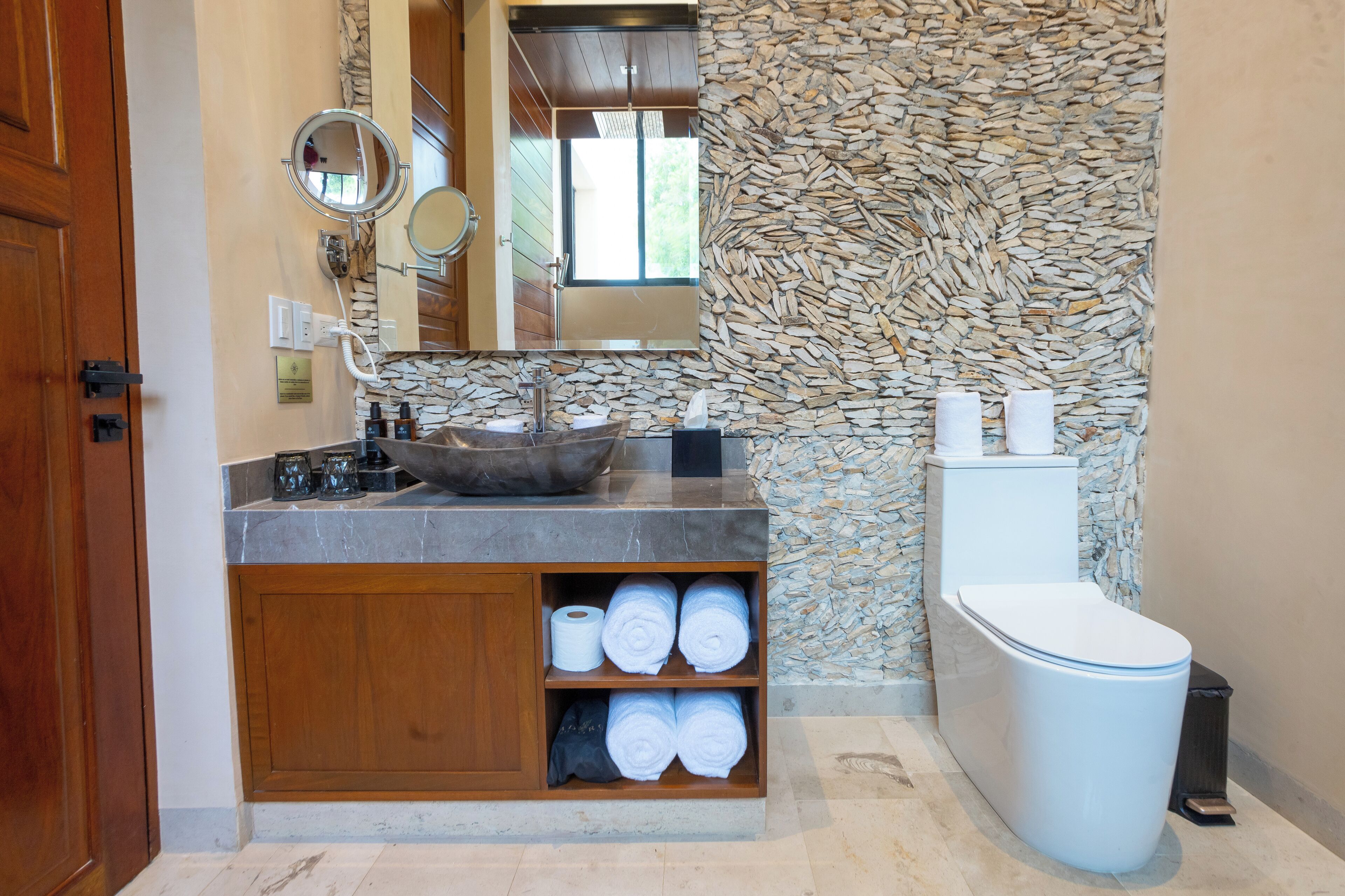 Une salle de bain avec un mur en pierre, un meuble-lavabo en bois, un lavabo en forme de cuvette et des toilettes.