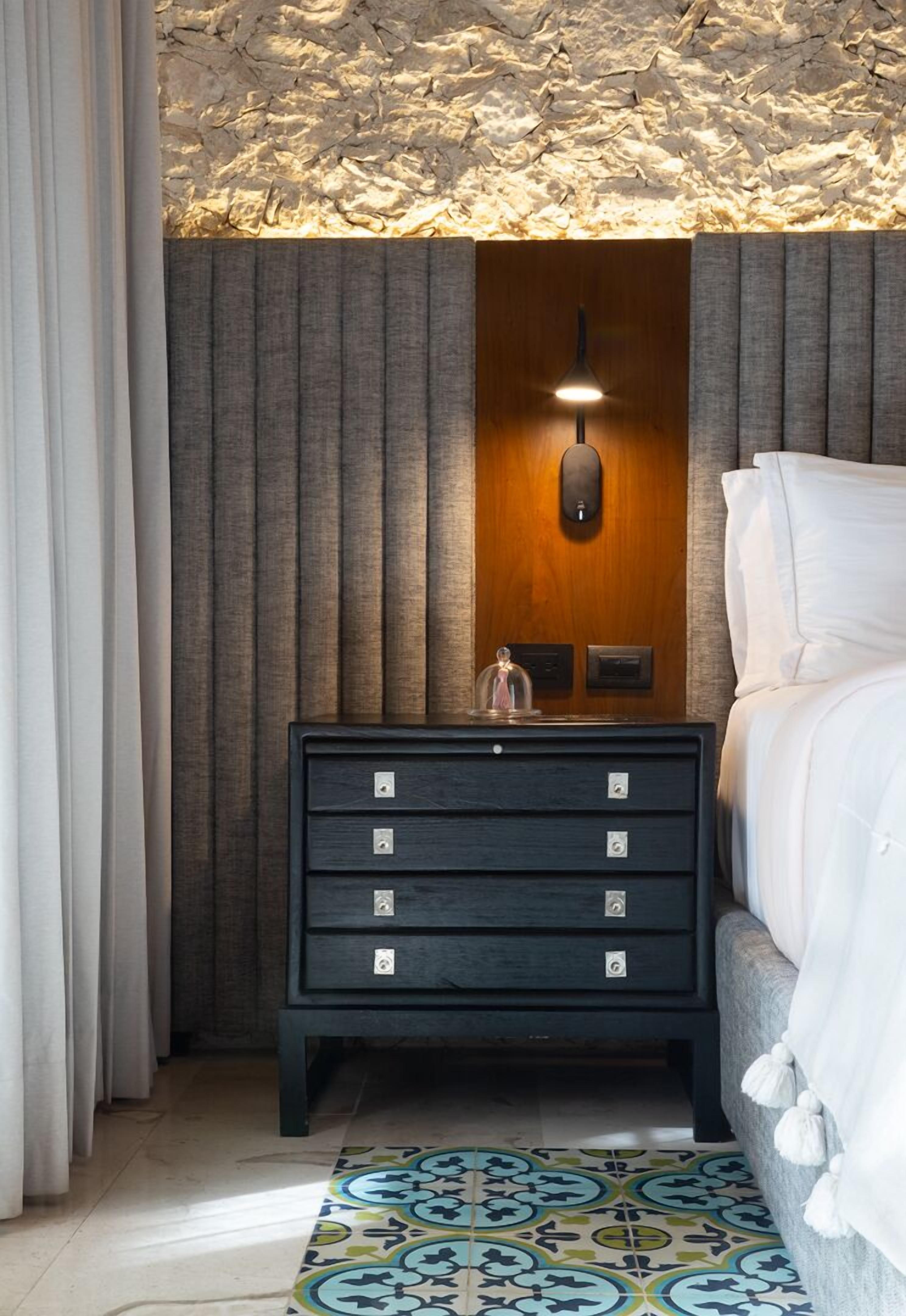 A bedroom with a bed, a nightstand with drawers, a wall-mounted light, and a patterned rug.