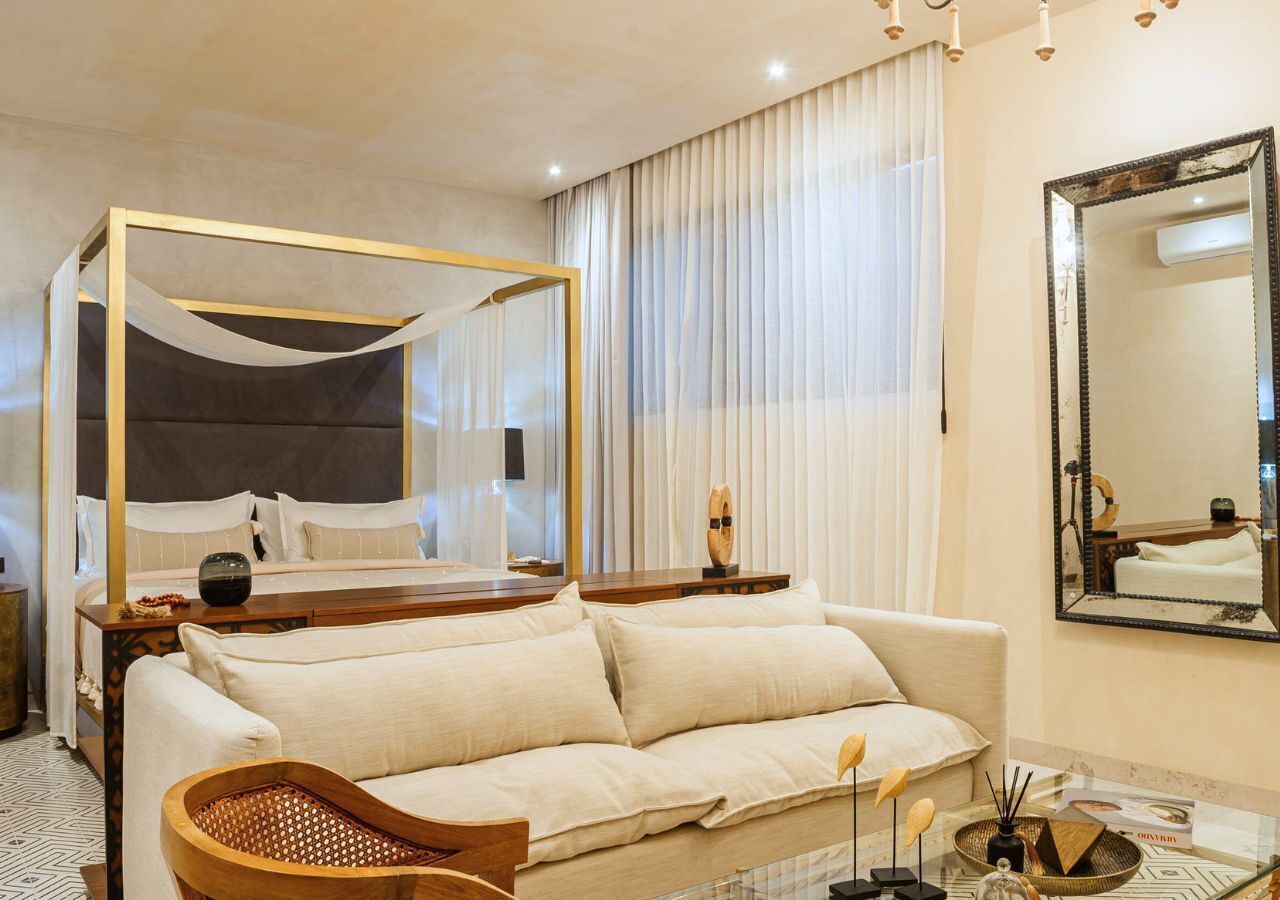 A modern hotel room with a four-poster bed, a sofa, a coffee table, and a large mirror.