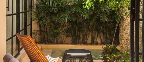Une terrasse ensoleillée avec une chaise en bois, une table ronde et une petite piscine, le tout entouré d’une végétation tropicale luxuriante.