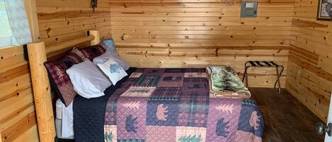 Une chambre dans une cabane en bois, avec un lit, des oreillers, une couverture, une fenêtre équipée d’un climatiseur et une petite étagère.