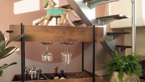 Un escalier moderne avec une rampe en verre, une sculpture équestre en bois, un chariot en métal contenant des bouteilles et des verres, et une plante en pot.
