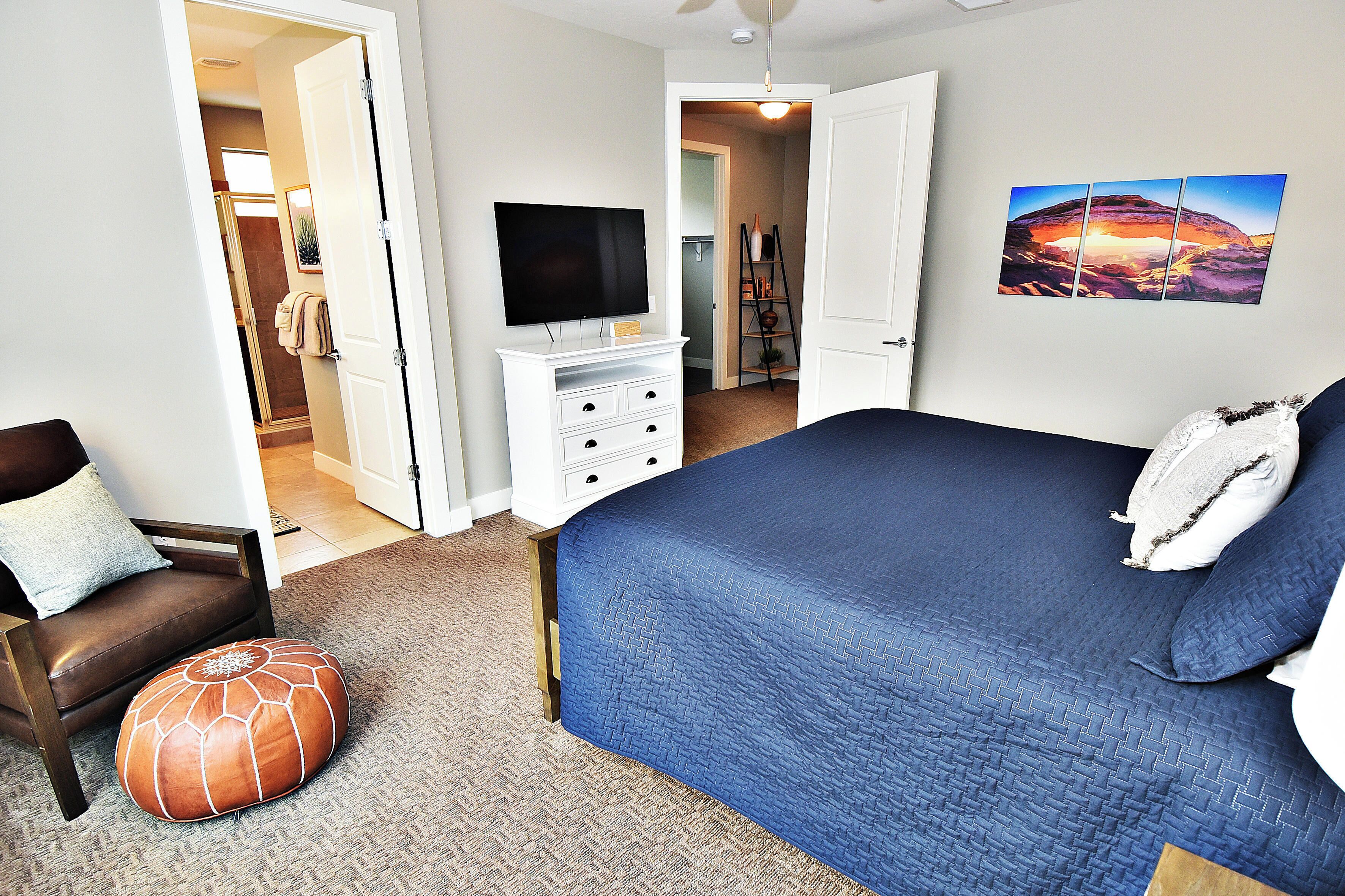 A bedroom with a large bed, a TV on a white dresser, a brown leather chair, and a door leading to a bathroom.