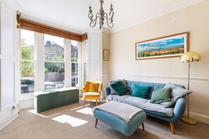A room with a blue sofa, a yellow armchair, a bench, a chandelier, and a painting on the wall. - Charming London Retreat (London)