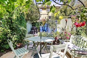 Een tuinpatio met een ronde tafel en stoelen, omgeven door weelderige begroeiing en potplanten.