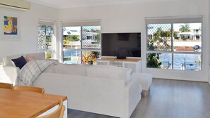 Una sala moderna con un sofá blanco, una mesa de centro de madera y un televisor de pantalla plana. Amplias ventanas ofrecen vistas al mar y un área exterior cercada.