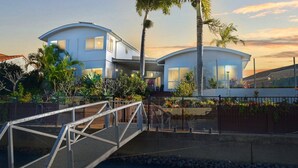 Una casa moderna de dos pisos con techo curvo, un puente y un jardín ajardinado con plantas tropicales.