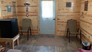 Une chambre de style rustique, avec des murs en rondins de bois, une porte en bois, deux chaises, une petite table, un téléviseur et une fenêtre avec des rideaux.