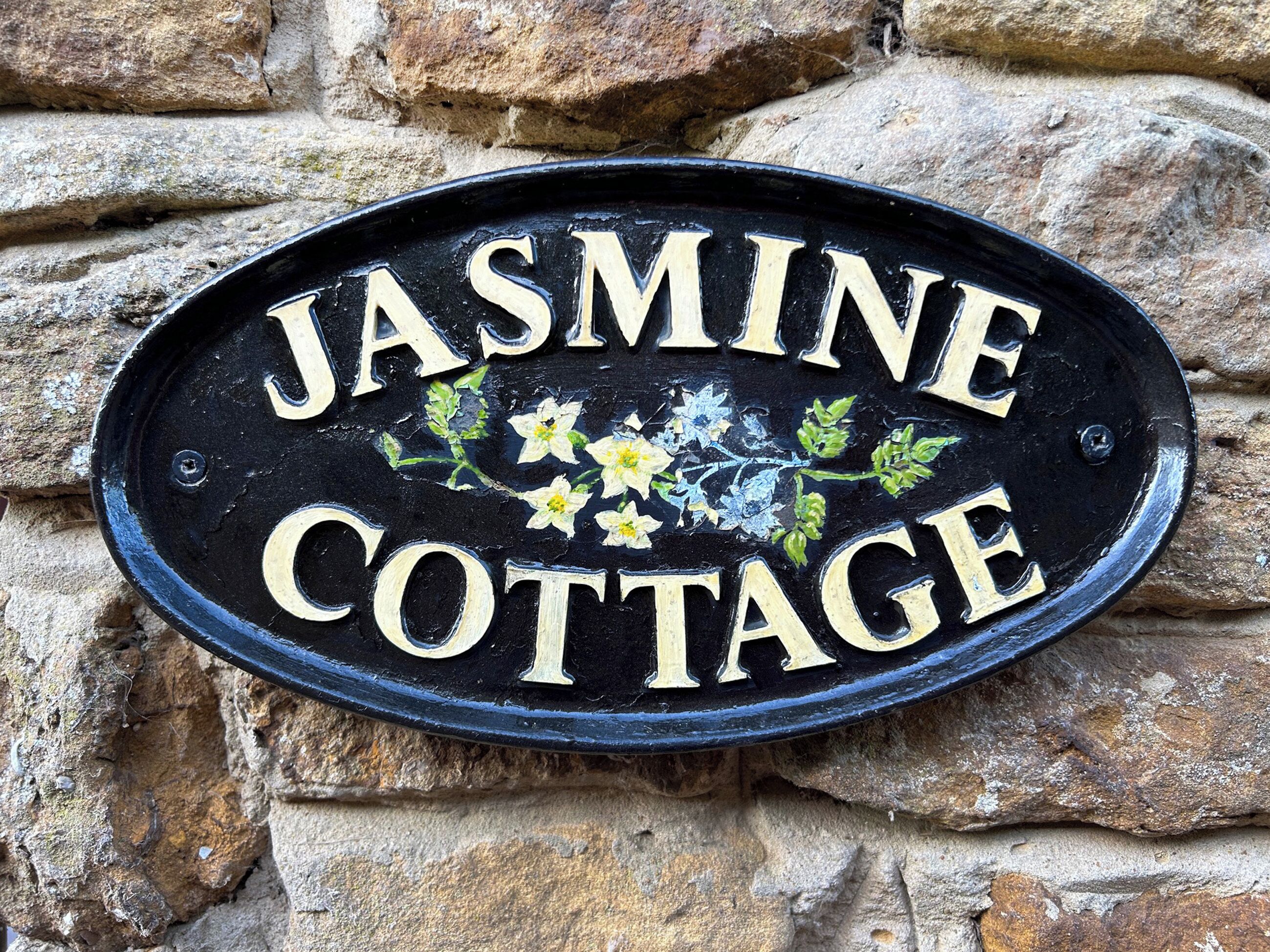 A black metal sign with the words "Jasmine Cottage" in white letters, surrounded by painted flowers and leaves.