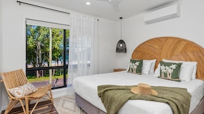 Une chambre avec un grand lit, une vue sur l’extérieur, un fauteuil en osier et un ventilateur de plafond.