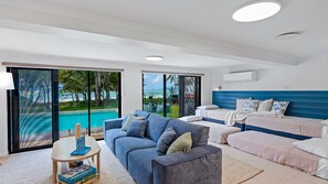 A modern hotel room with a large sliding glass door leading to a pool, a sofa, a bed, and a small table with books.