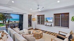 A spacious living room with a large sofa, a round wooden coffee table, and a view of the beach through an open door.
