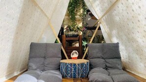 Ein gemütliches Zelt mit grauen Kissen und einem gewebten Hocker in der Mitte, umgeben von Holzdielen und einer Ziegelwand.
