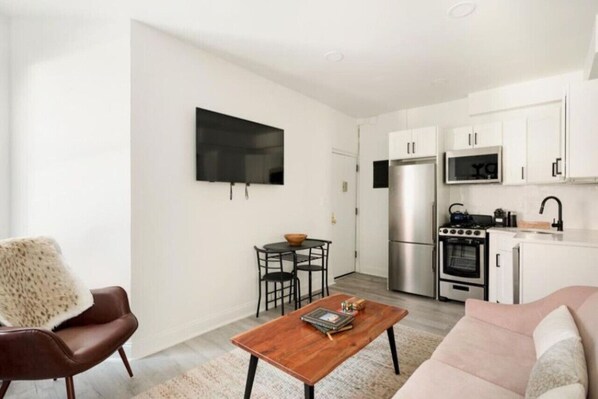 A compact living space with a kitchenette, a flat-screen TV mounted on the wall, a dining area with a table and chairs, and a sofa.