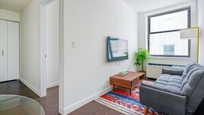 A modern living room with a grey sofa, a small wooden coffee table, a TV mounted on the wall, a potted plant, and a large window with a view of the city. - 2 Bedroom Gem in Financial (New York)